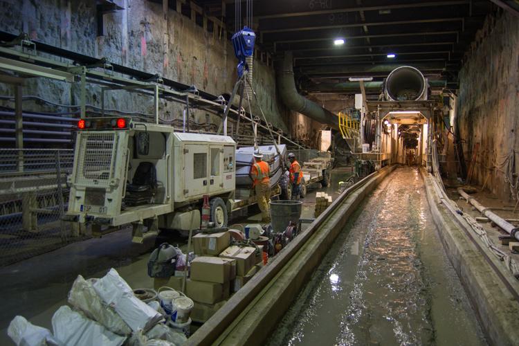 Workers in hard hats operate tunnel boring machine equipment in underground subway construction tunnel with water channel.
