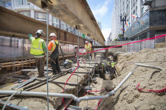Construction workers in safety gear at Central Subway project site with excavation equipment and pink safety barriers.