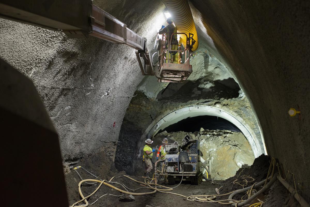 Workers operate equipment inside tunnel excavation with barrel vault structure and exposed rock walls.