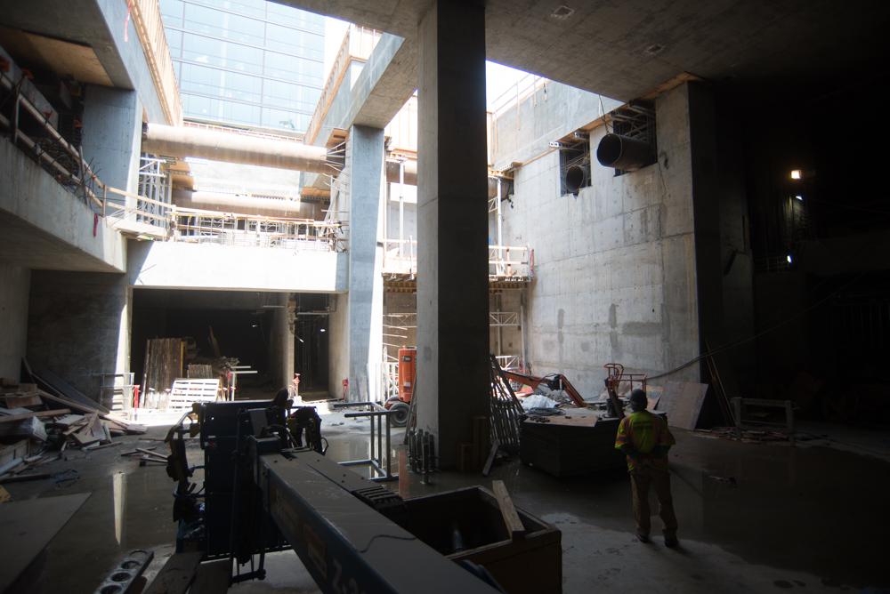 Construction site interior showing concrete pillars, exposed walls, and workers in safety gear beneath skylights.