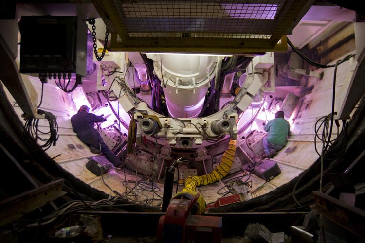 Workers perform welding and assembly inside the trailing gear section of a tunnel boring machine, illuminated by purple work lights.