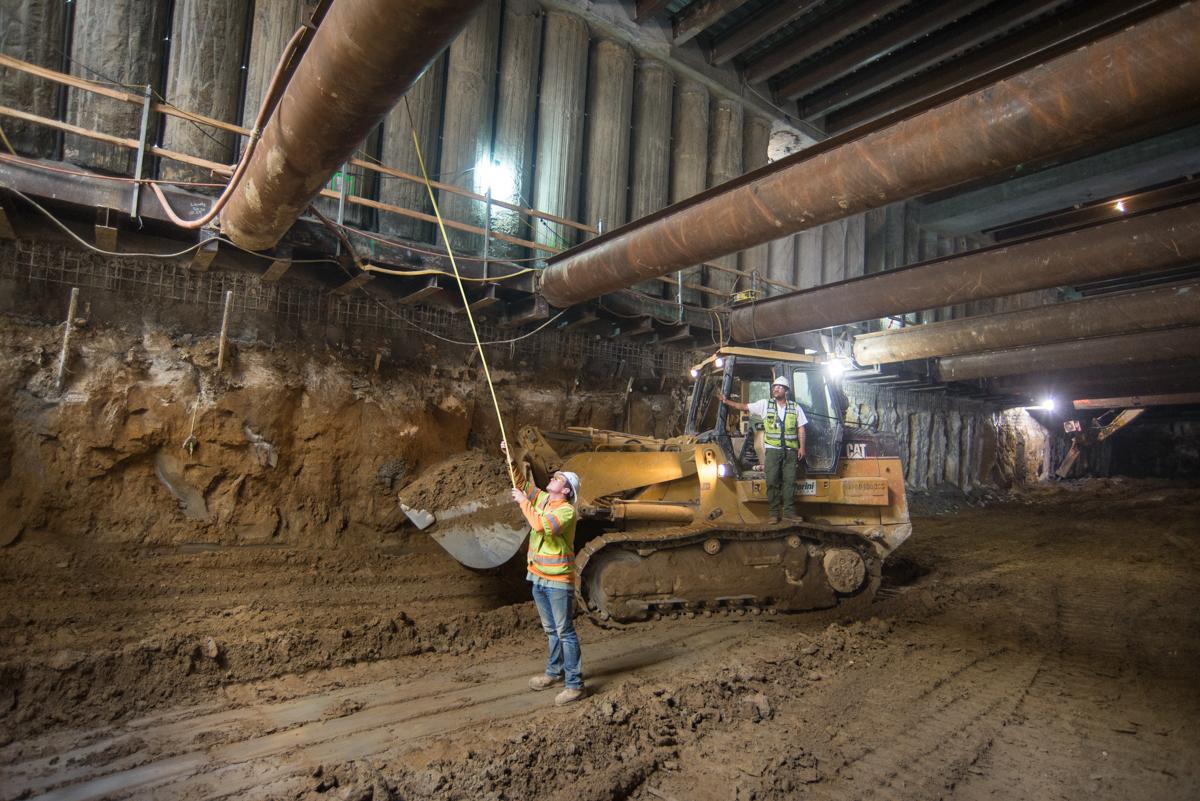 Worker in safety vest guides steel bucket suspended by crane in underground station excavation site.