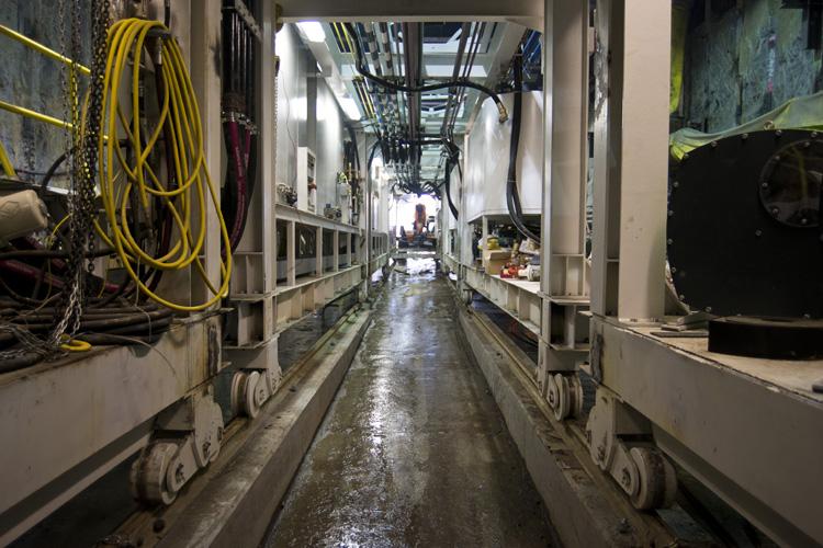 Interior view of tunnel boring machine trailing gear with yellow cables and equipment along narrow metal corridor.