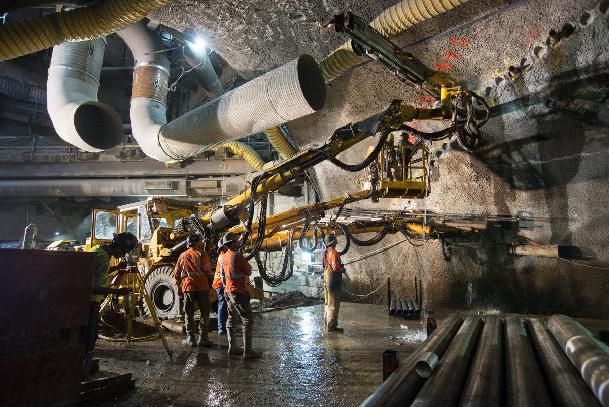 Workers in orange vests operate a tunnel boring machine drill rig underground at a subway construction site.