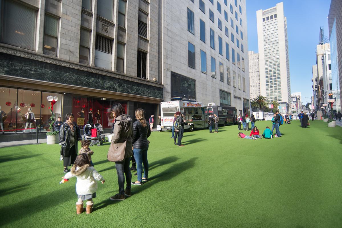 Families and children enjoy a green lawn plaza with food trucks near downtown San Francisco buildings.