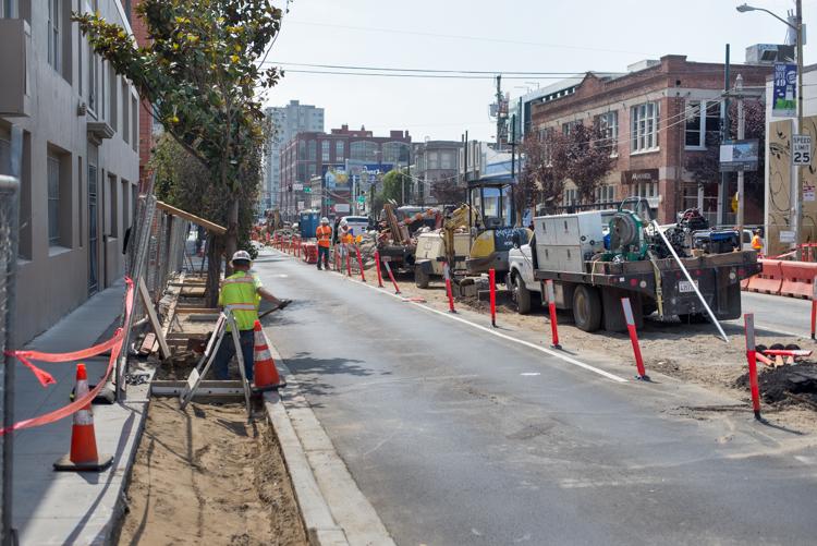 Construction work on 4th Street with equipment, workers in safety vests, red barriers, and surrounding urban buildings.