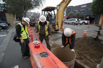 Construction workers install utility infrastructure and OCS pole foundations on 4th Street near active excavation equipment.
