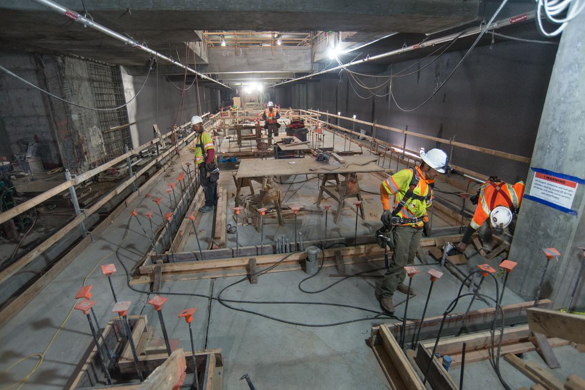 Workers in safety gear construct interior walls and rebar cages inside an underground transit station headhouse.