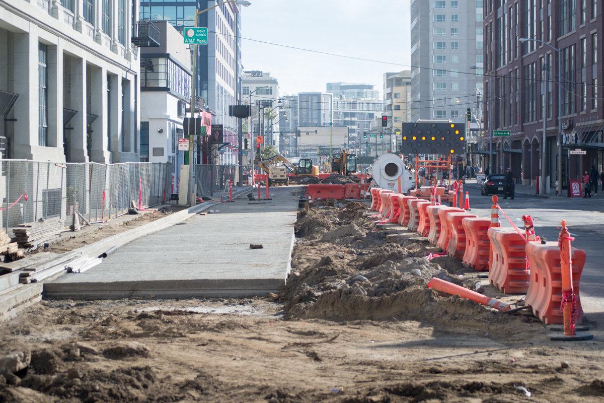 Urban street under construction with excavation, concrete work, and red safety barriers lining the project area.