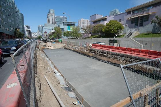 Construction site with freshly poured concrete slab on 4th Street, surrounded by chain-link fencing and equipment, with city skyline in background.