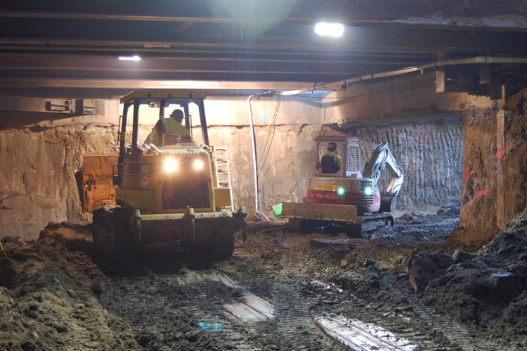 Excavation equipment and workers in underground tunnel construction site with concrete walls and overhead lighting.