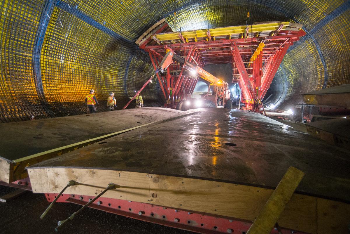 Workers construct concrete forms inside a tunnel under yellow and blue safety netting with red support structures.