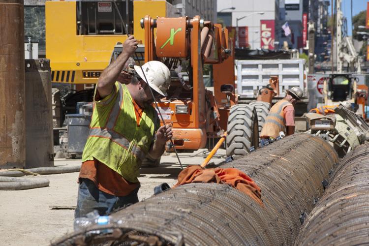 Construction worker in safety vest and hard hat operating equipment near large reinforced concrete cylinders on urban street.