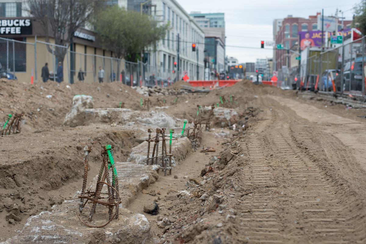 Construction excavation site with exposed foundation piles marked with green flags along urban street.