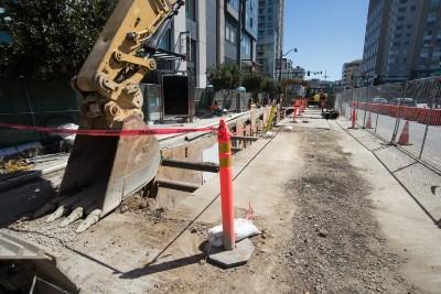 Construction site with excavator and red barriers along urban street trench for sewer line upgrade work.