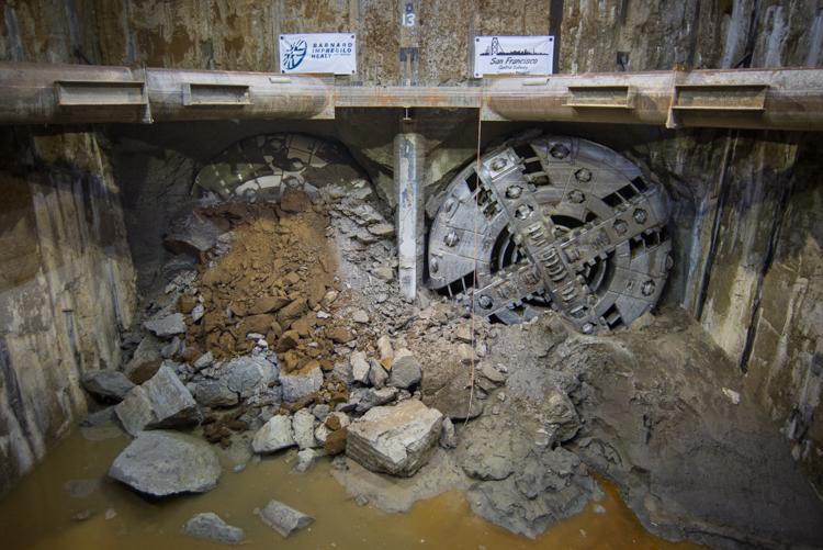 Tunnel boring machine cutter heads inside underground excavation with debris and water pooling at base.