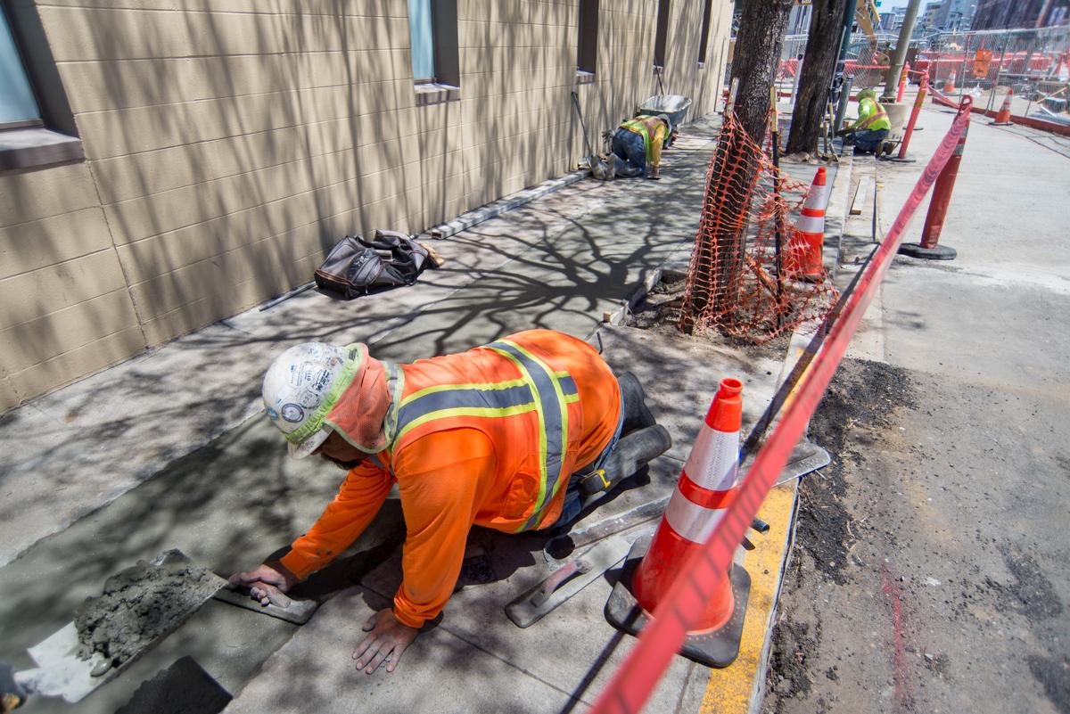 Construction worker in orange safety vest smoothing concrete on sidewalk during Central Subway project work.