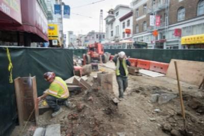 Construction workers in safety vests excavating underground transit station shaft in urban street surrounded by protective barriers and equipment.