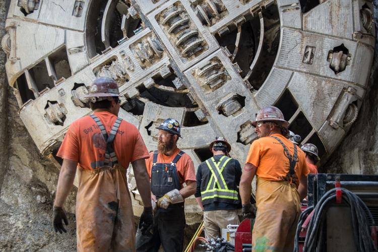 Construction workers in safety gear stand before a large tunnel boring machine during dismantling operations.