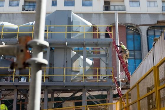 Construction workers install ventilation system on Chinatown Station headhouse with yellow safety scaffolding and equipment visible.