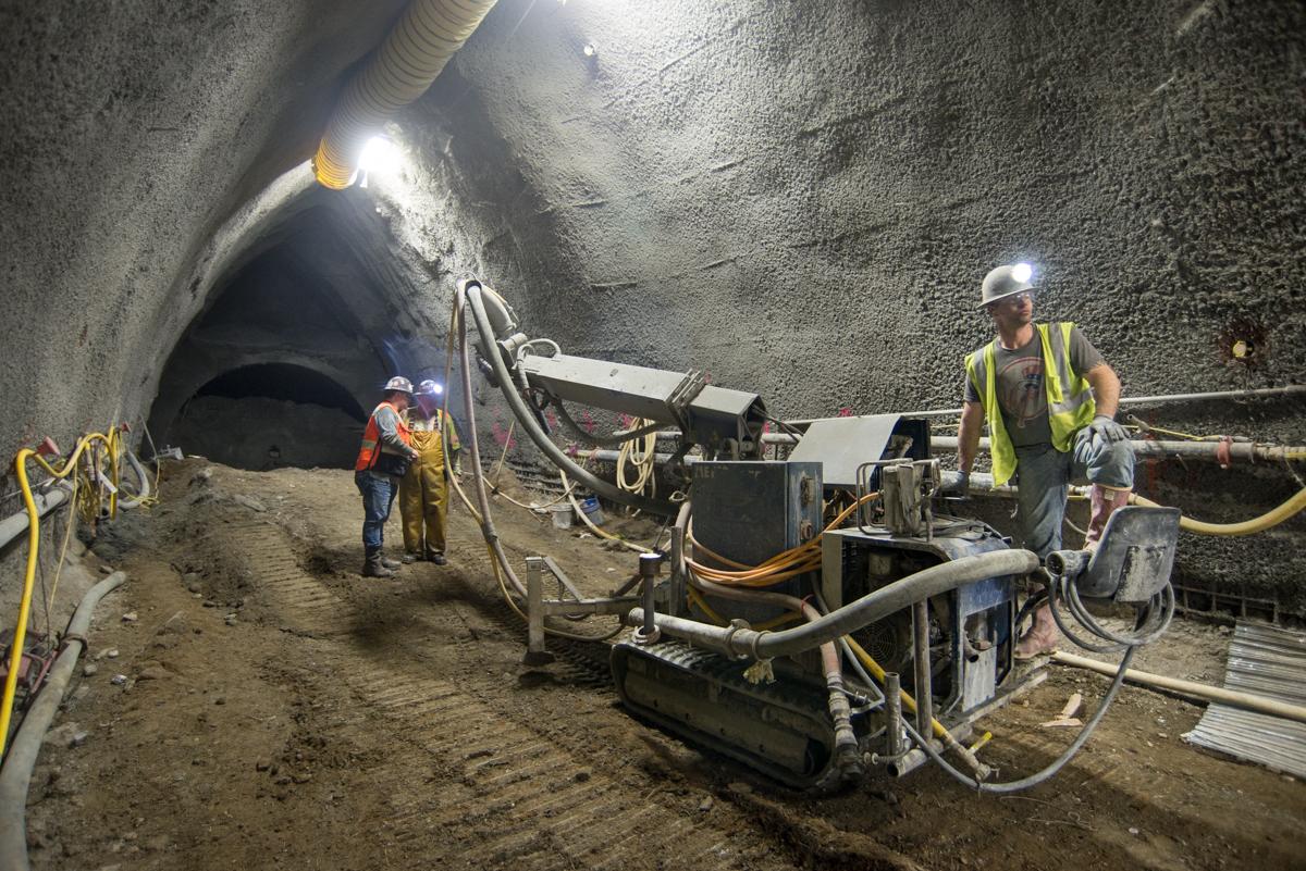 Workers operate drilling equipment inside subway tunnel excavation site with exposed rock walls and overhead lighting.