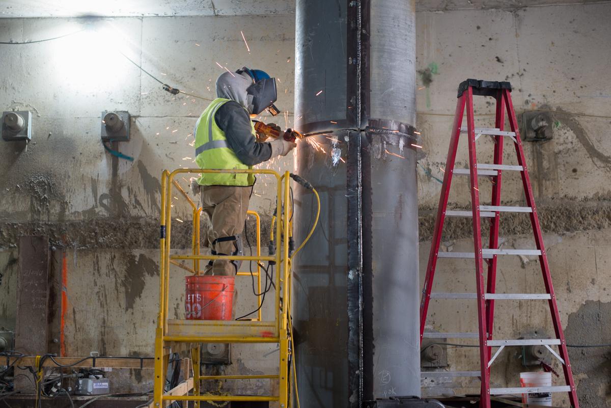 Worker in yellow lift welding steel bracing around support column in underground transit tunnel.
