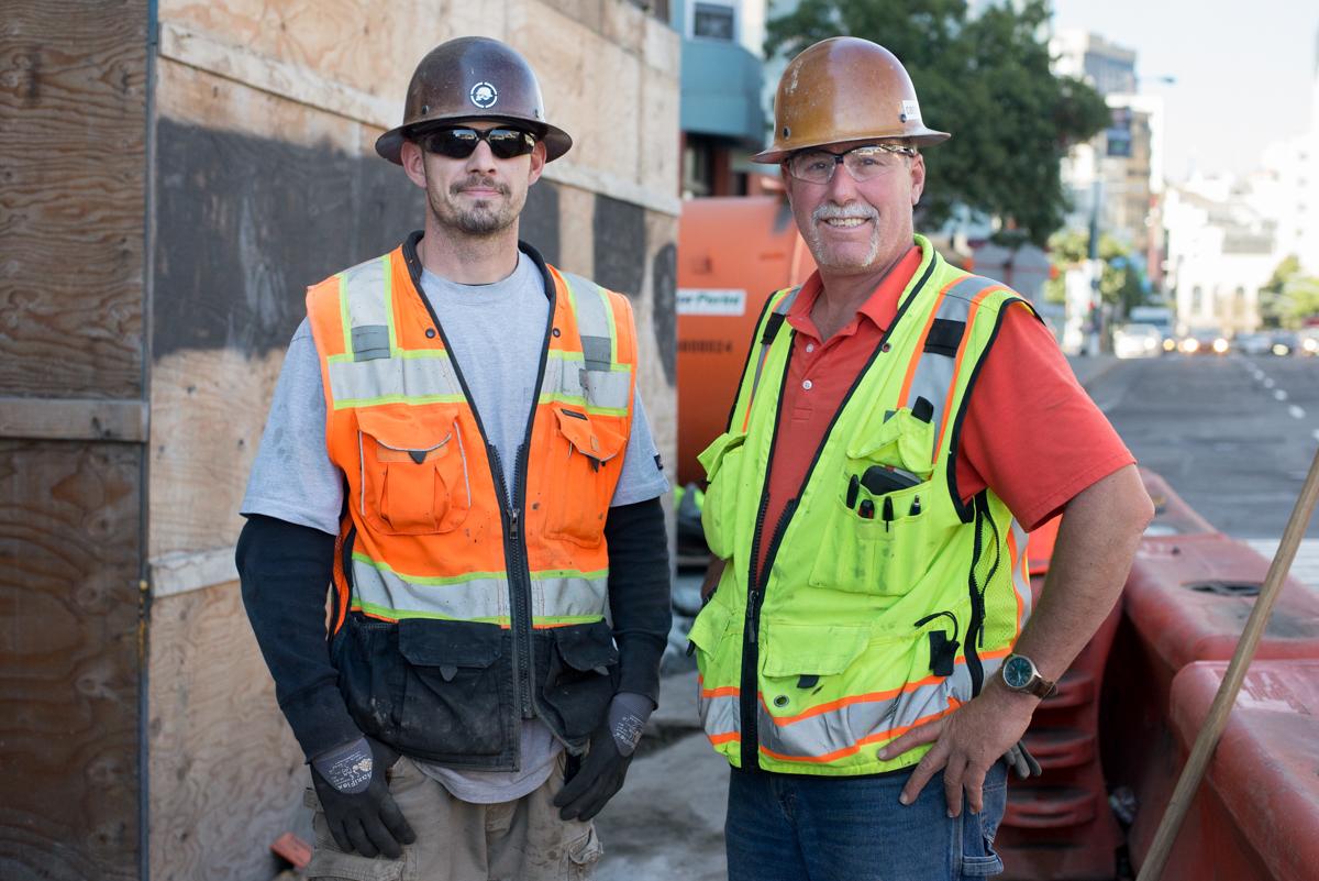 Two construction workers in safety vests and hard hats stand on a job site.