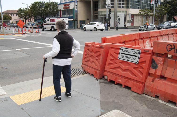 Older pedestrian with cane navigates around orange construction barriers on city sidewalk.