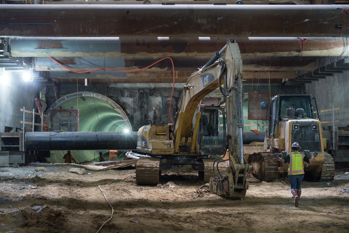 Construction crew operates excavator and compactor equipment inside underground station structure with steel bracing visible.