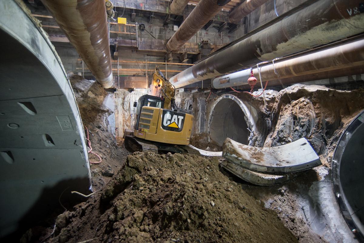 CAT excavator removing soil from subway tunnel construction site with large pipes overhead.