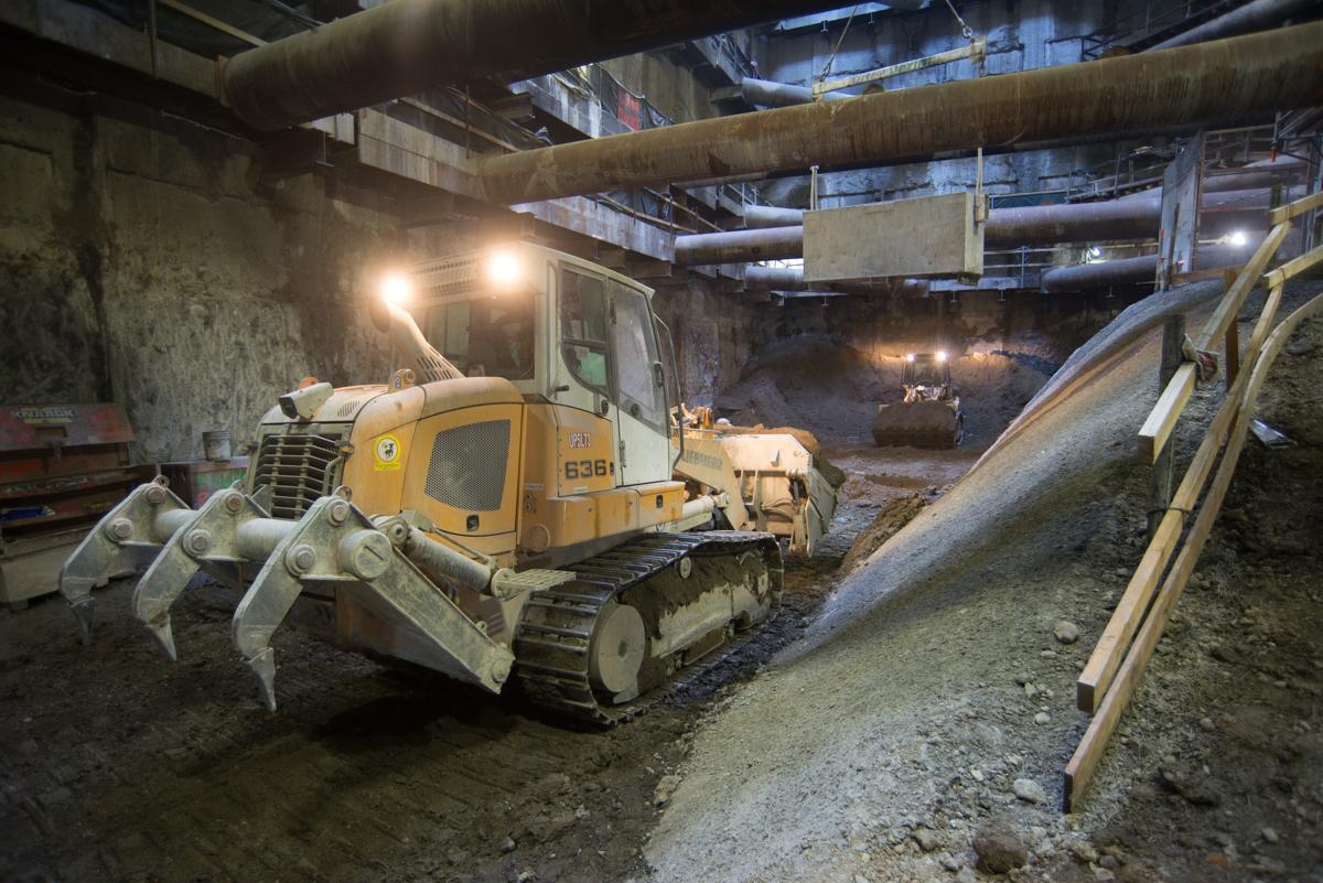 Yellow tracked excavator with clamshell bucket in underground construction site with pipes and equipment overhead.