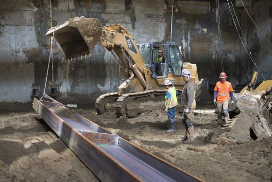 Excavator positioning steel beam inside transit station headhouse with construction workers nearby.