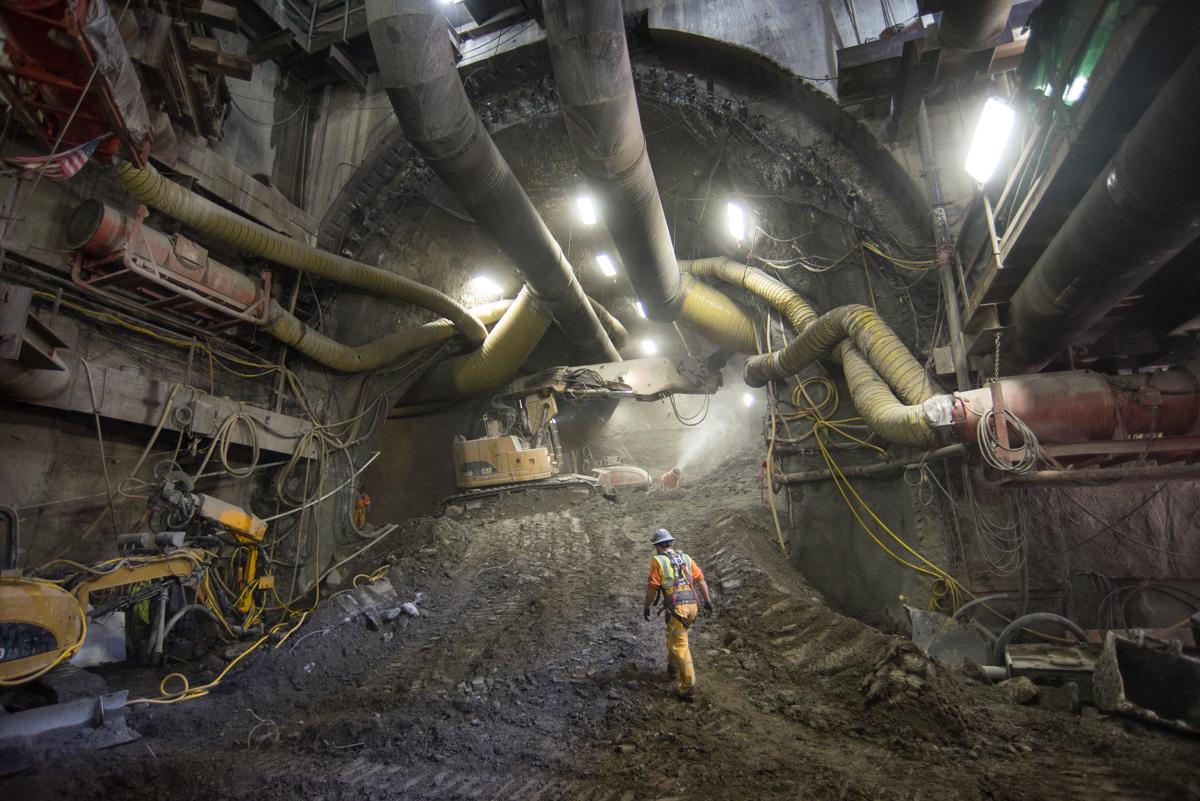 Worker in hard hat and safety vest inside underground tunnel with large pipes and construction equipment.