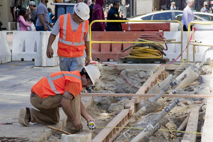 Construction workers in safety vests and hard hats work on underground utility lines at a street excavation site with pedestrians and traffic visible in background.