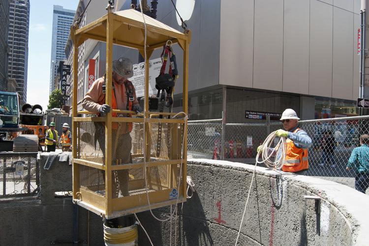 Construction worker descending into Ellis Street shaft in yellow safety cage during ground improvement work.
