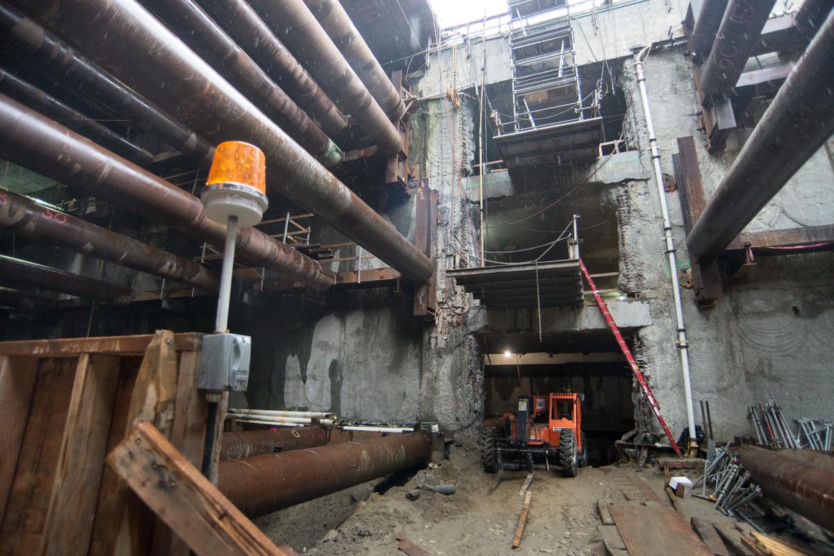 Construction site with large pipes, concrete truck, orange warning light, and metal scaffolding in underground station excavation.