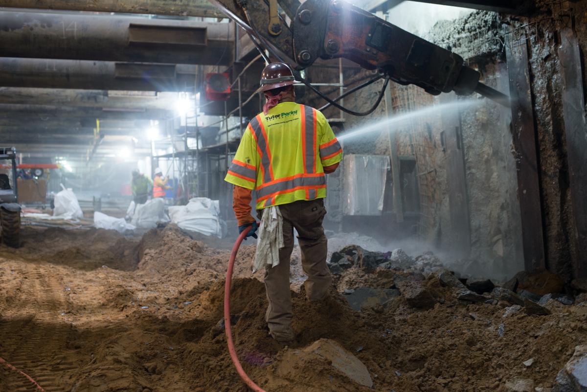 Worker in safety gear sprays water while operating jackhammer on concrete wall at underground construction site.