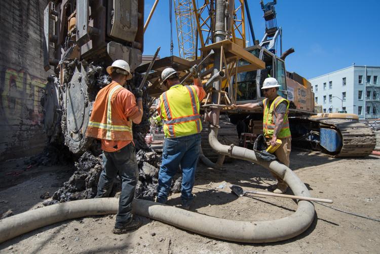 Three workers in safety gear maintain a hydromill machine at a construction site.