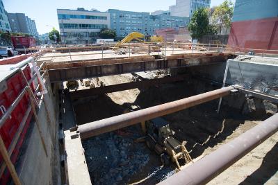 Underground construction site with excavated pit, temporary steel support beams, and surrounding urban buildings.