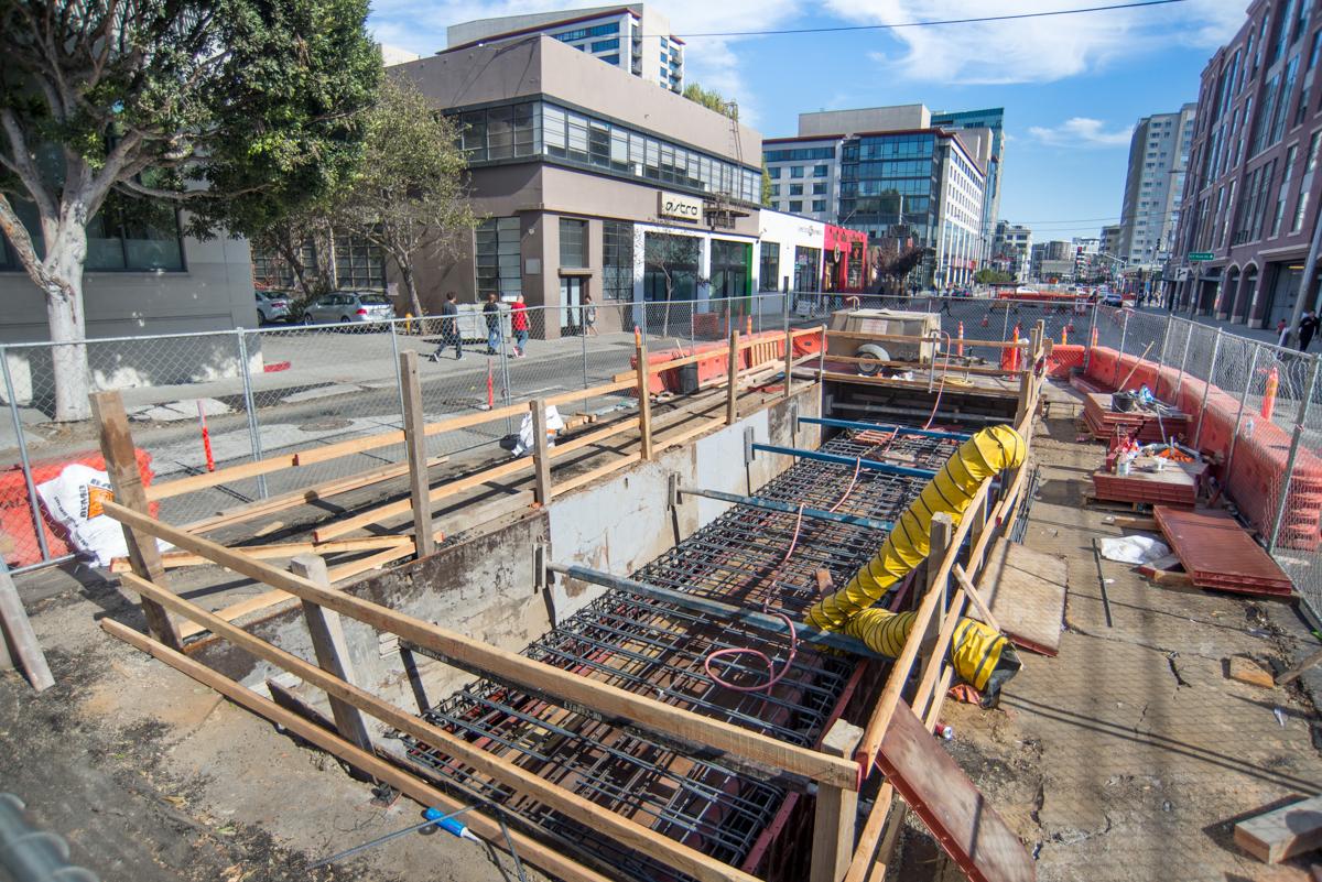 Construction site showing underground sewer line work with wooden barriers, yellow equipment, and metal reinforcement on urban street.