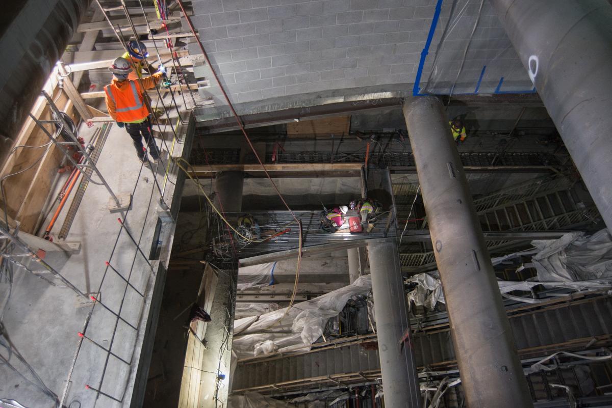 Worker in orange safety vest on scaffolding above underground transit station under construction with exposed concrete, steel framing, and electrical systems.