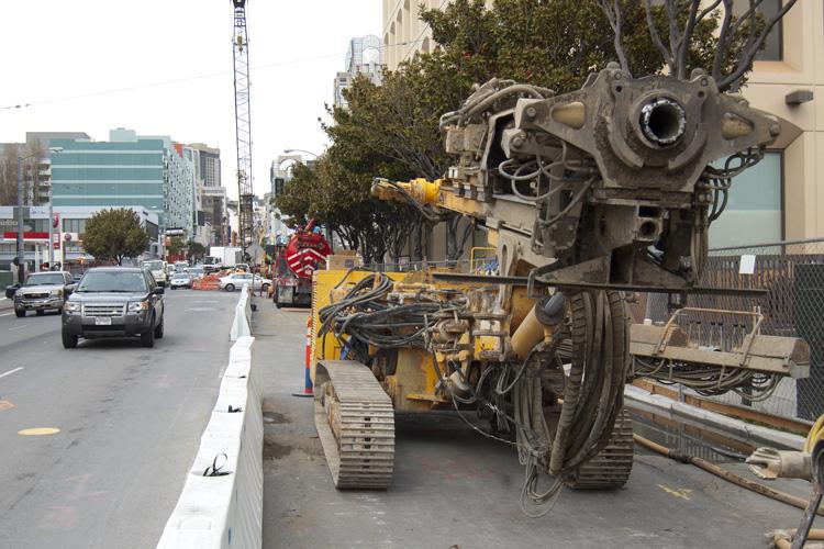 Heavy construction equipment with articulated arm on 4th Street work site, with traffic and urban buildings visible.