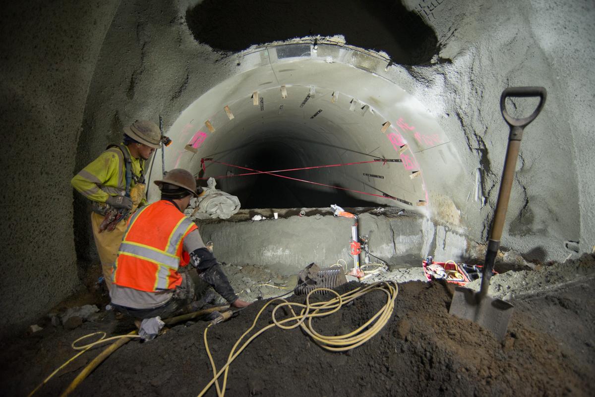Workers in safety gear excavating inside a large circular tunnel with concrete barrel vault rings during station cavern construction.