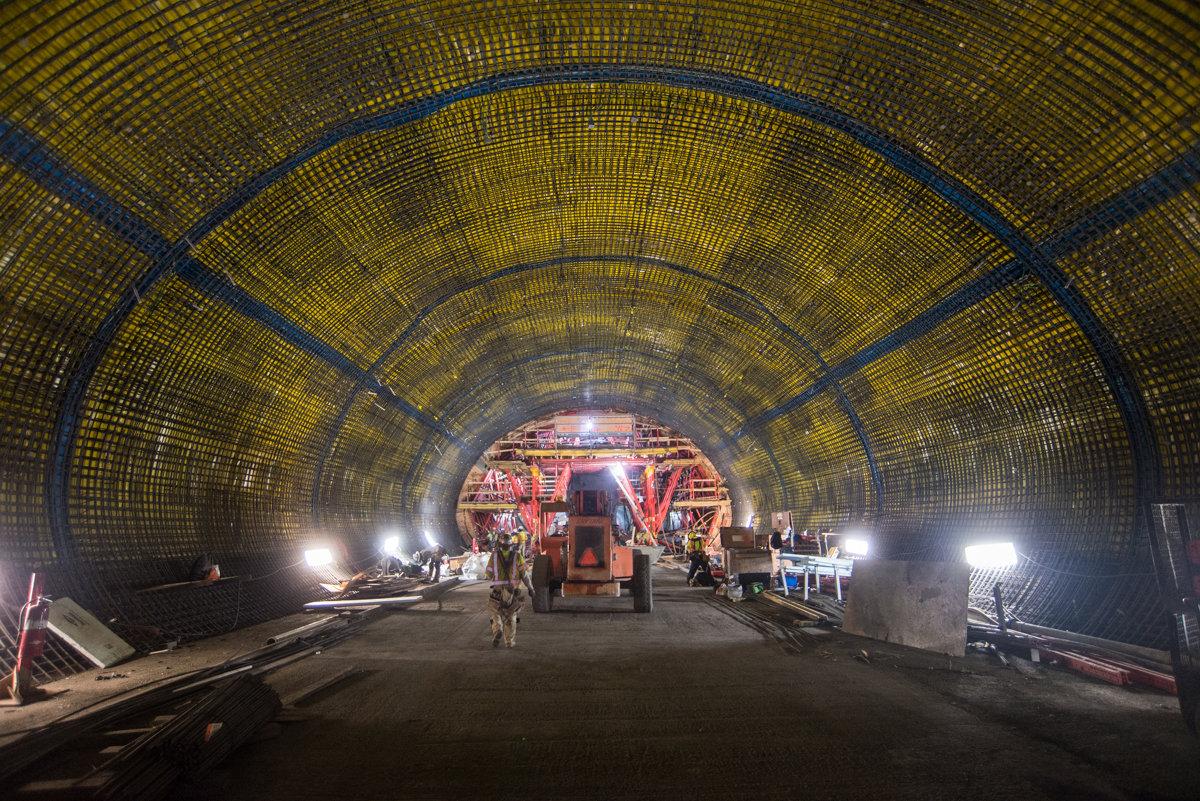 Construction workers and equipment inside illuminated tunnel with curved yellow mesh walls and blue support beams.