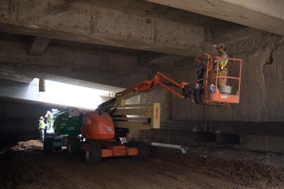 Worker in elevated bucket inspects concrete structural elements during underground excavation beneath transit headhouse roof slab.