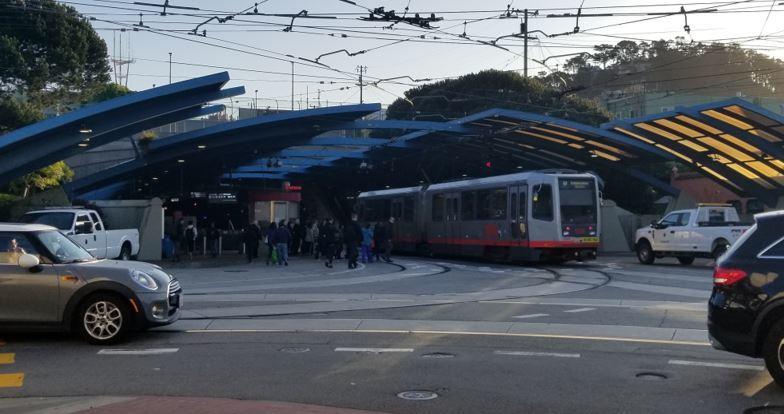 Light rail train at West Portal station with pedestrians, cars, and overhead power lines visible.
