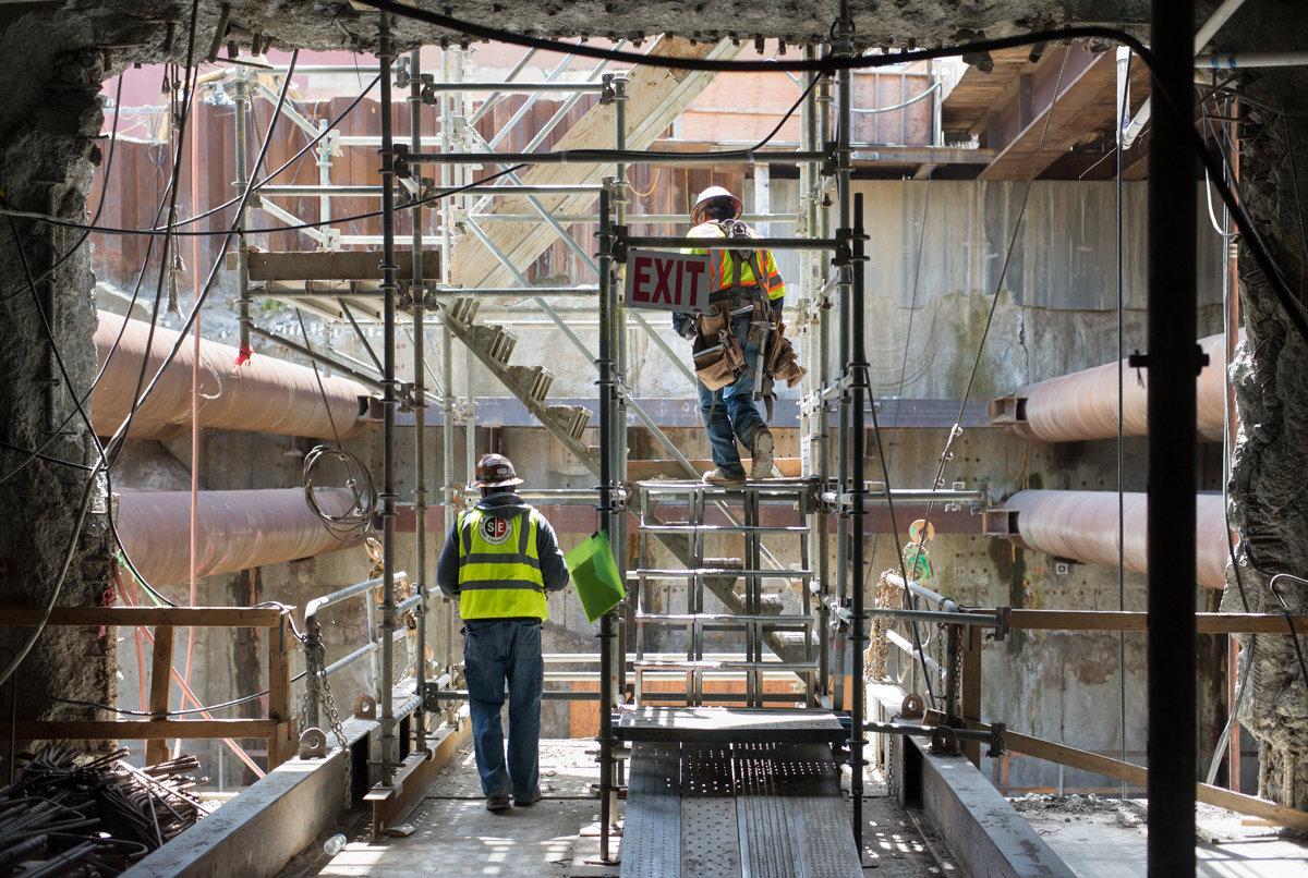 Two construction workers on scaffolding inside a transit station undergoing renovation work.