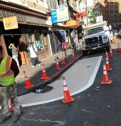 Construction crew installing gray pedestrian safety zone markings on 6th Street with traffic cones and barriers.