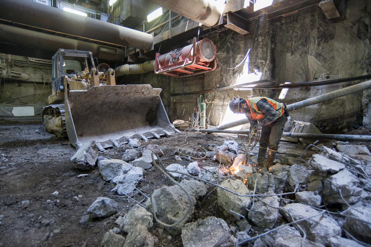 Worker operating jackhammer on concrete rubble inside underground tunnel construction site with excavator and equipment visible.