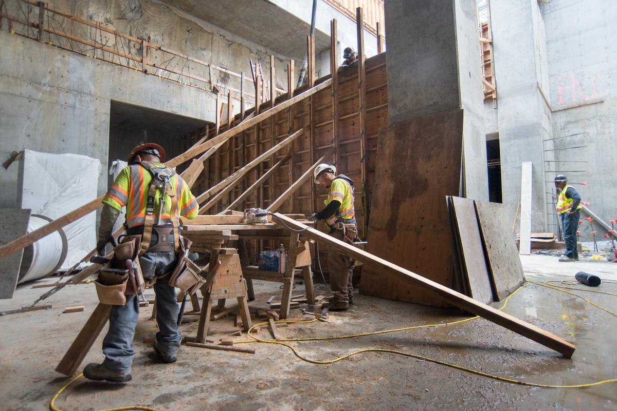 Construction workers in safety vests build wooden formwork for escalator support walls inside a concrete transit station.